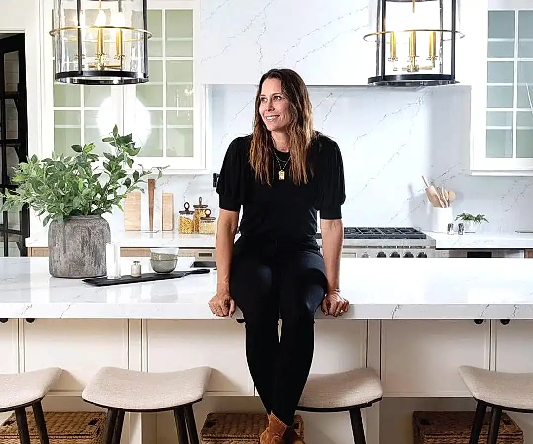 Siyah kıyafetli bir kadın, modern beyaz dolaplar ve sarkıt ışıklarla çevrili bir mutfak adasında oturuyor ve bir iç mimarın vizyonunun zarafetini temsil ediyor. Tezgahı süsleyen bir bitki ve mutfak eşyaları, sofistike ancak işlevsel tasarıma katkıda bulunuyor.