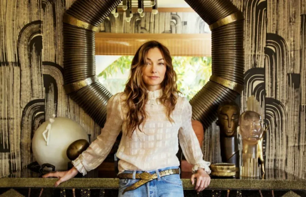 Kelly Wearstler uzun saçlarıyla, beyaz bir bluz ve kot pantolonla, dekoratif bir duvar aynasının önünde duruyor. Eklektik dekor, zevkli bir şekilde tasarlanmış odaya çekicilik katan benzersiz heykeller ve desenli duvar kağıtlarıyla bu ünlü yabancı kadın mimarın yaratıcı vizyonunu andırıyor.