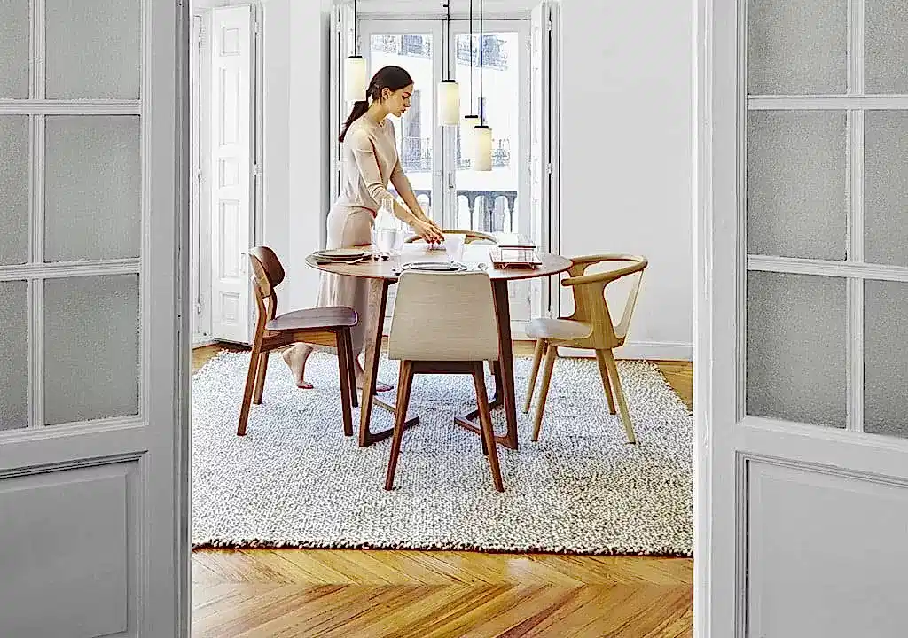 Bir kadın, iç mimarların tasarımlarını andıran, parke zeminli ve yüksek pencereli aydınlık bir odada, ahşap sandalyelerle donatılmış yuvarlak bir yemek masası düzenliyor.