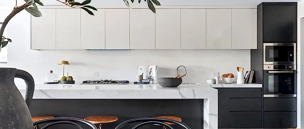 Beyaz dolaplar, siyah aletler, tabureli mermer ada ve bir kase limon bulunan modern mutfak. Tezgahı süsleyen dekoratif eşyalar ve bir bitki, profesyonel bir iç mimarın dokunuşunu anımsatan şık bir hava yayıyor.