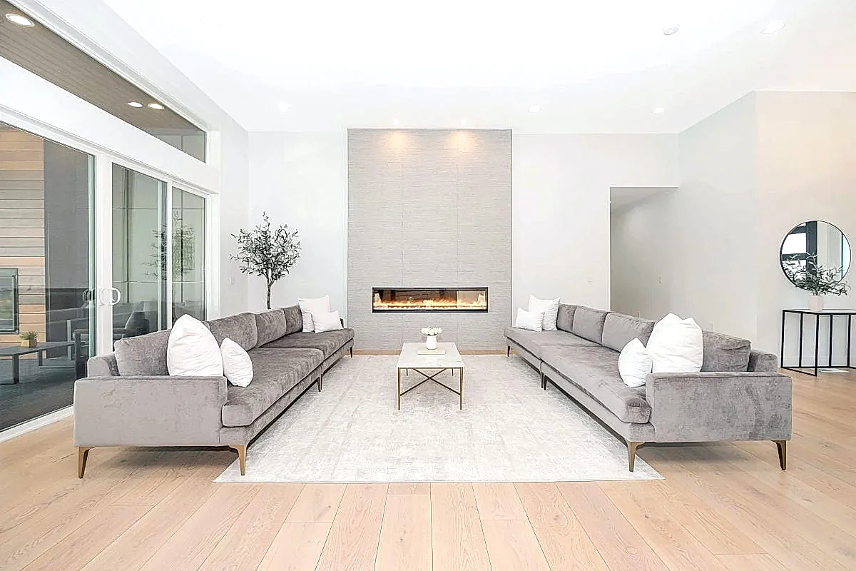 Bir iç mimar tarafından tasarlanmış, birbirine bakan iki gri kanepe ve ortada bir sehpa bulunan modern bir oturma odası. Alan, arka duvardaki doğrusal bir şömine ve açık renkli ahşap döşeme ile zenginleştirilmiş olup, her villa veya çağdaş tasarım için mükemmel olan kusursuz bir stil karışımı sergiliyor.